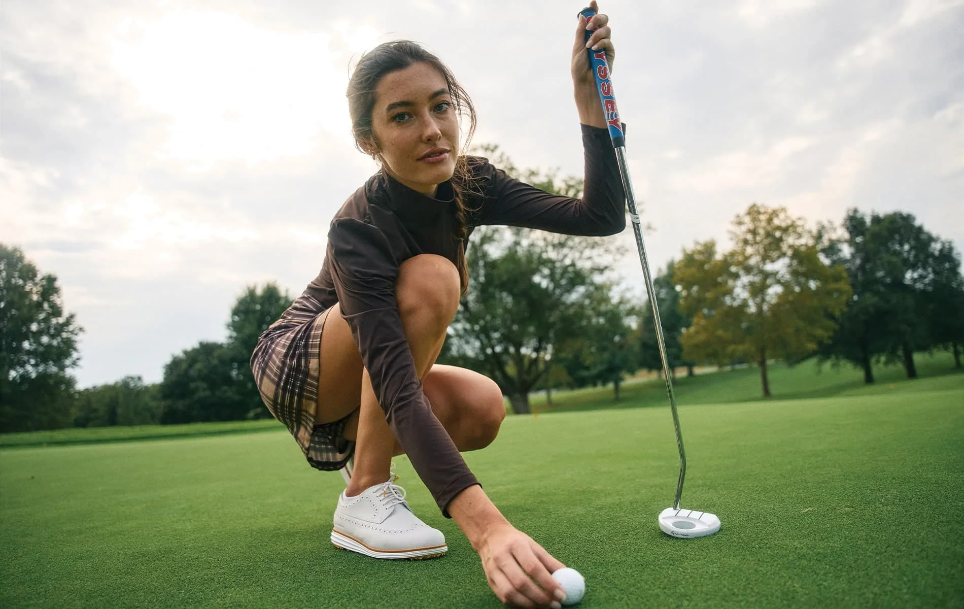 A golfer in a brown mock-neck top and plaid skirt crouching to place a ball on a putting green, a putter upright in her other hand.