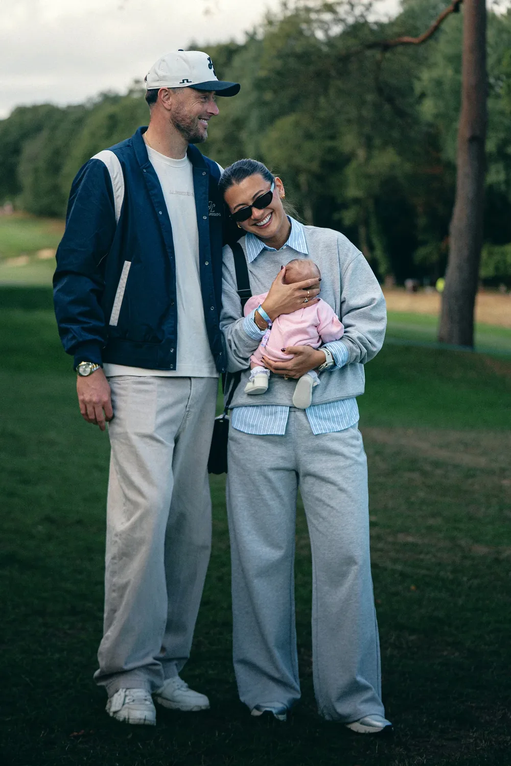 A couple in relaxed J.Lindeberg layers standing on a golf course, the woman holding a baby.