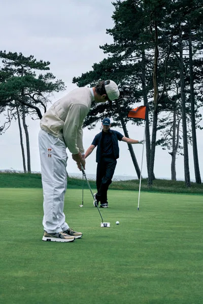A golfer in a light grey J.Lindeberg outfit putting on a green lined with pine trees.