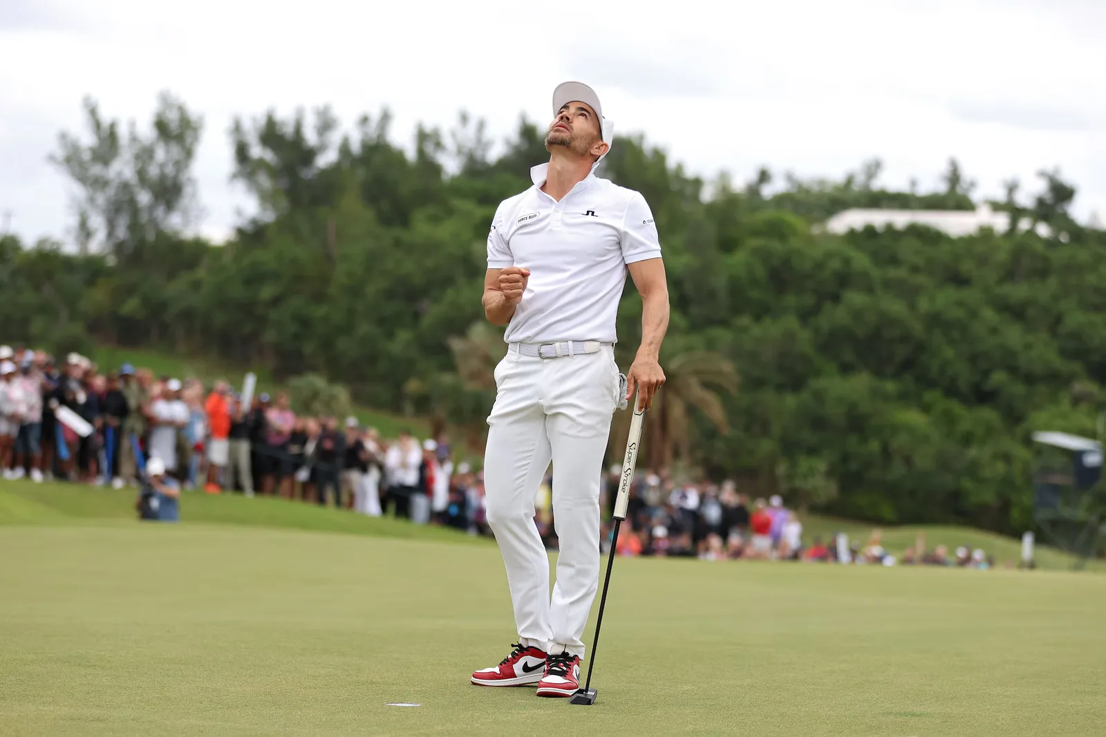 Camilo Villegas in white standing on a green watching a putt, with a gallery behind him.