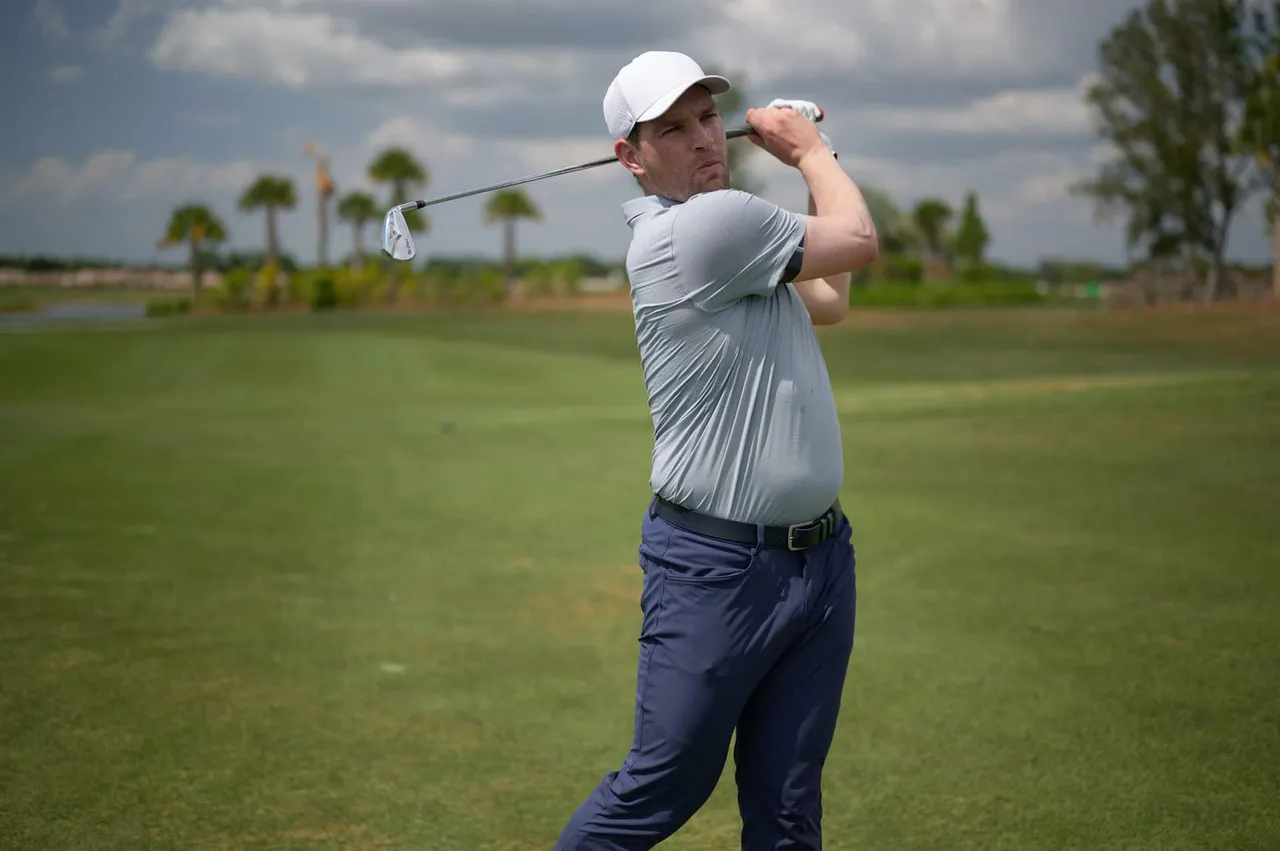 A golfer in a light blue polo and white cap holding his finish on a range lined with palm trees.