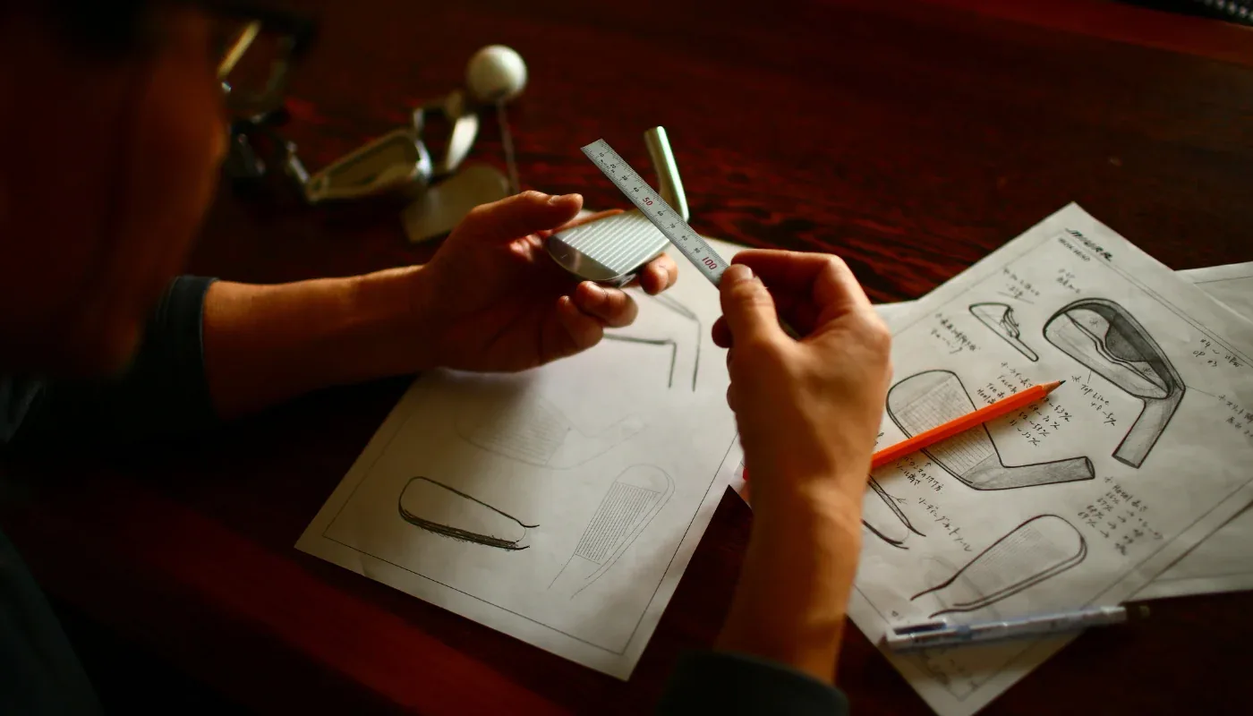 Hands measuring an iron head over hand-drawn design sketches.