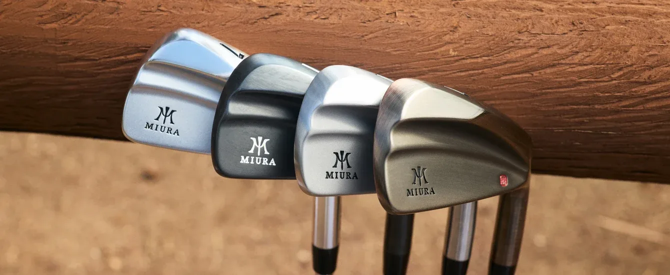 Four Miura irons in different finishes resting on a wooden rail.