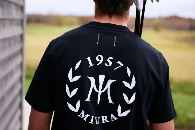 A golfer seen from behind in a black T-shirt printed with a laurel logo reading 1957 Miura, carrying clubs.