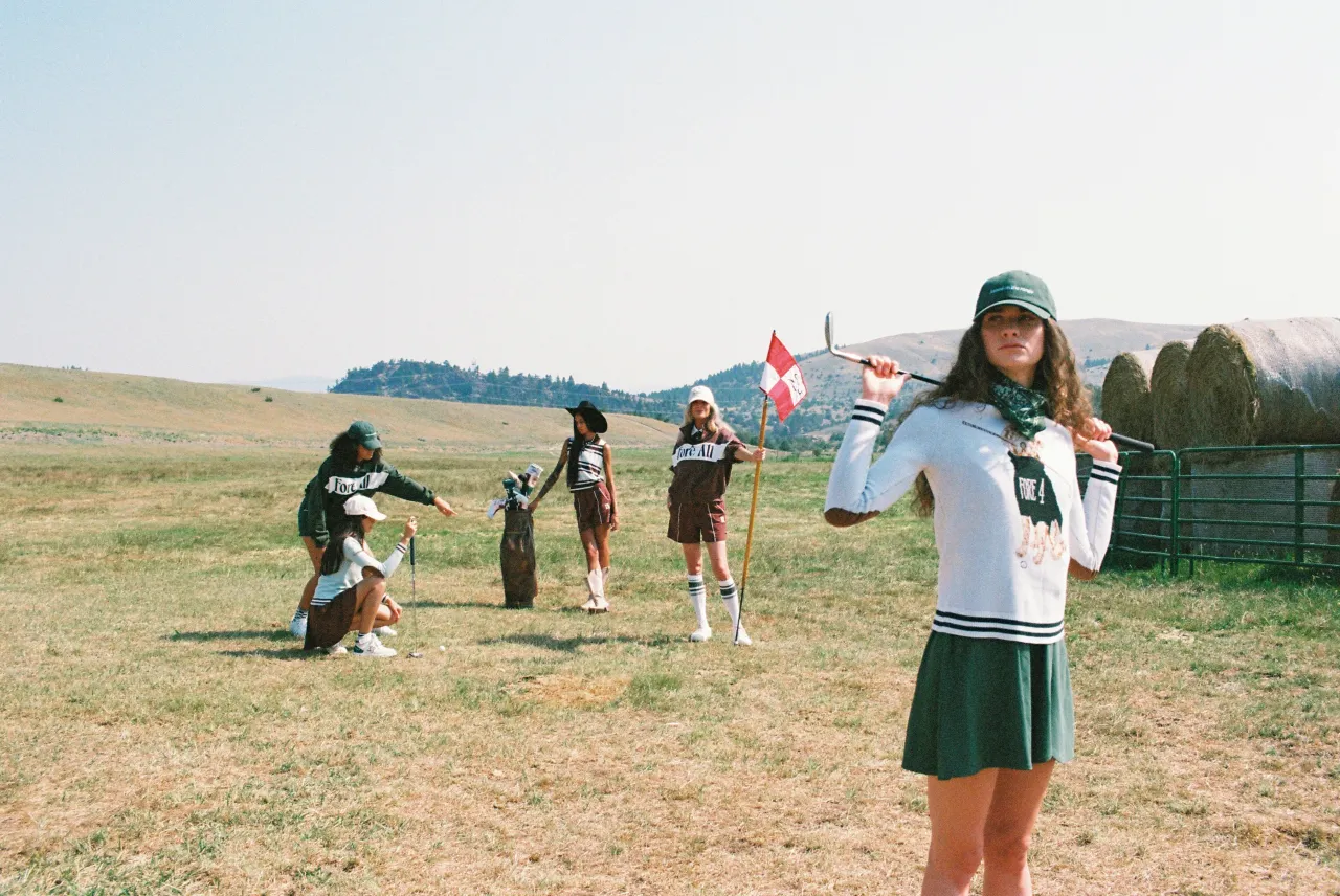 A woman in a white sweater and green pleated skirt holding a club across her shoulders in a grass field, with friends gathered around a flagstick behind her.