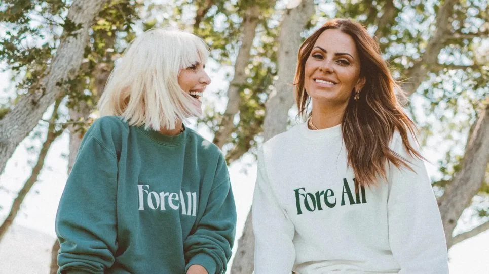 Fore All co-founders Jen Clyde, left, in a green Fore All sweatshirt, and Michelle Money, right, in a white one, laughing outdoors.
