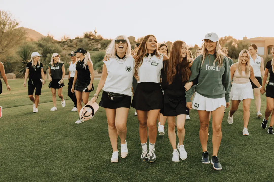 A group of women walking arm in arm down a fairway, several of them in Fore All apparel.