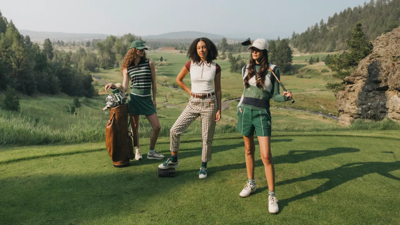 Three women on a tee box above a pine-lined valley in Fore All golf apparel, one beside a leather carry bag and one with a club over her shoulder.