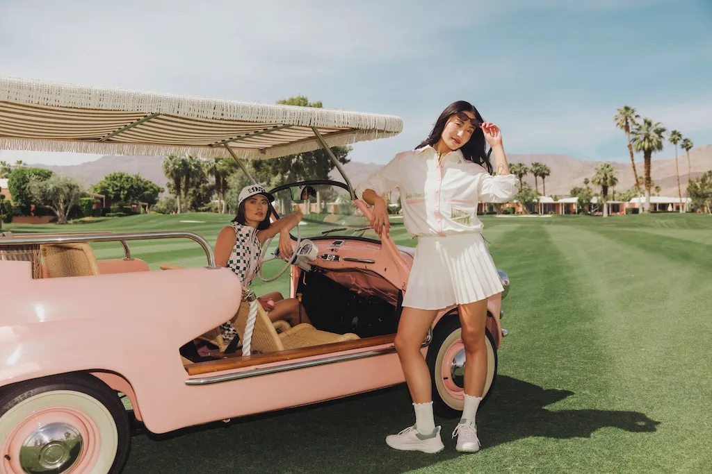 Two women in white golf outfits beside a pink vintage cart on a desert course lined with palm trees.