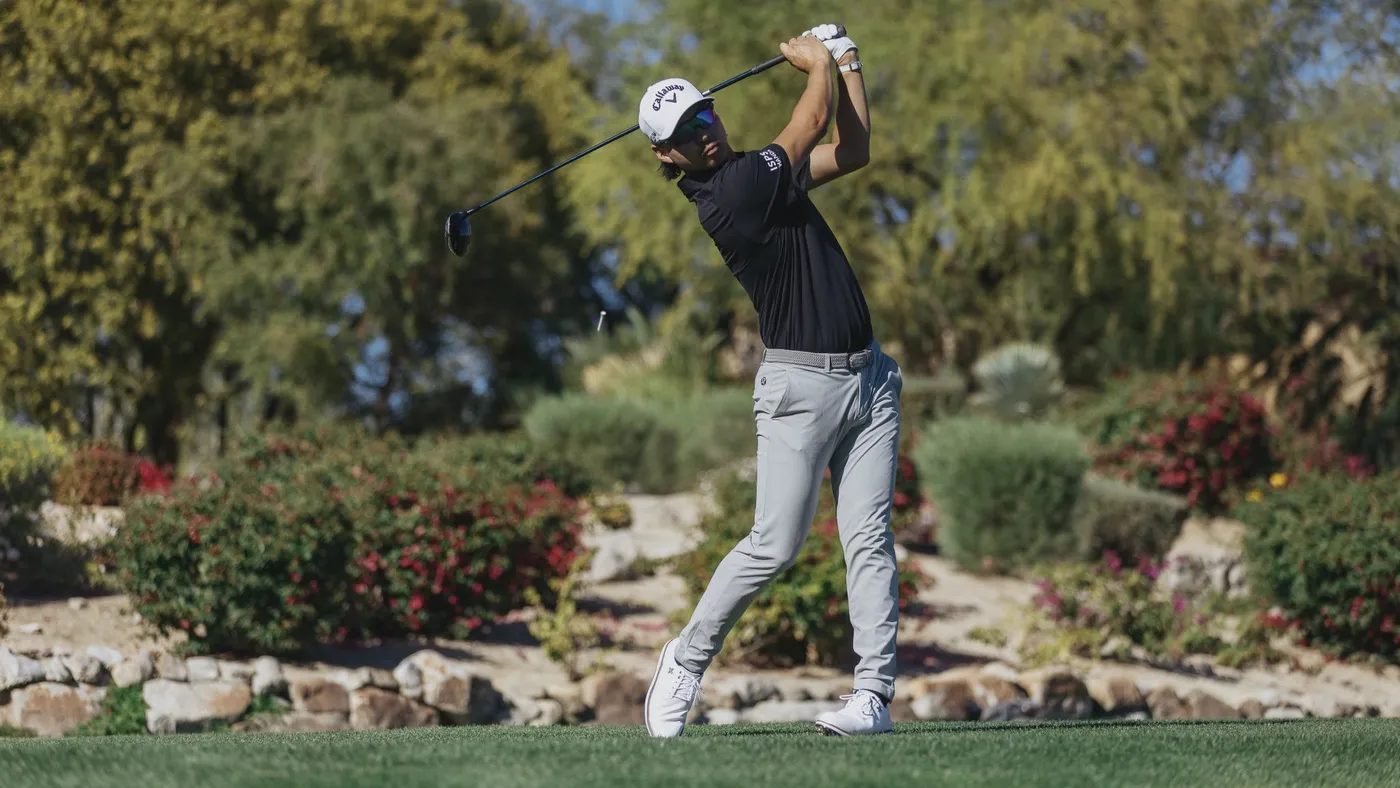 Min Woo Lee holding his finish after a drive in white golf shoes and grey trousers.