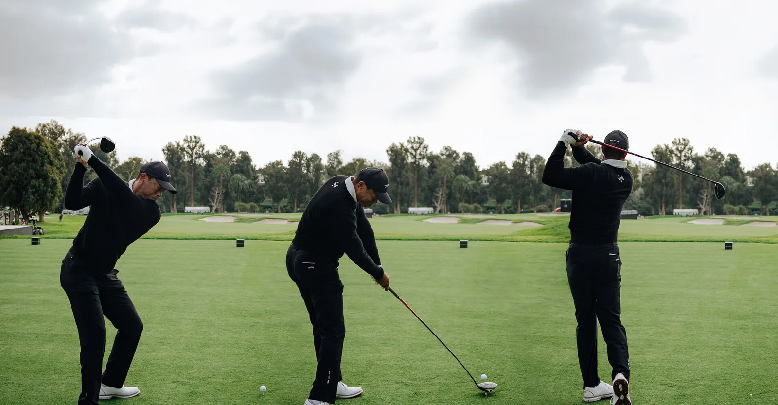 Three golfers dressed in black swinging on a practice range.