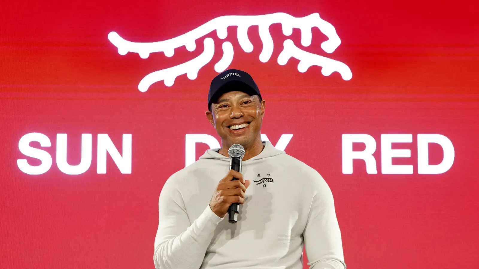 Tiger Woods speaking on stage in front of a red Sun Day Red backdrop showing the tiger logo.